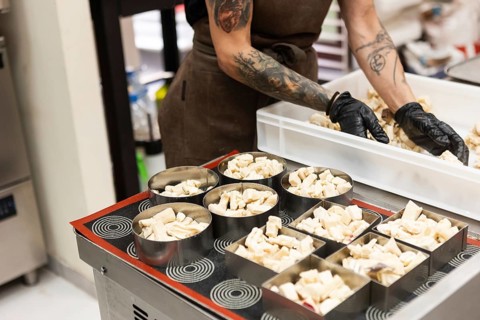 A focused baker with tattooed arms and black gloves arranges pastry pieces into metal molds,