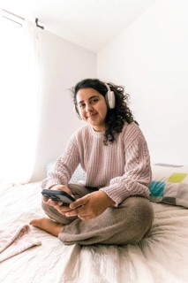 A young latin woman enjoying leisure time at home, wearing headphones, and using a smartphone while