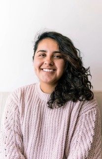 A young latin woman with curly hair smiles warmly, sitting indoors wearing a cozy sweater The