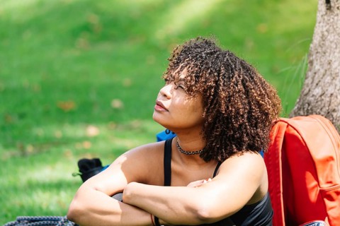 A Black woman sits under a tree in a Bogota park, resting her arms on her knees while gazing into