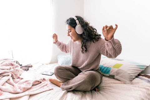 A young latin woman sits cross-legged on her bed, wearing headphones and a cozy sweater She appears