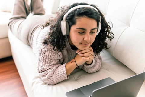 A young latin woman relaxes on a couch, wearing headphones and a cozy sweater, as she engages with