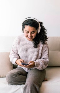 A young latin woman sits comfortably on a couch, wearing headphones and looking at her phone She