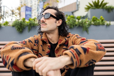 A fashionable man wearing sunglasses and a patterned jacket sits on a bench outdoors, exuding a