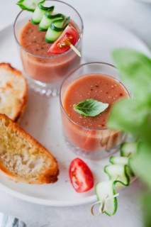 Two glasses of gazpacho topped with cucumber and tomato skewers, alongside toasted bread, served on