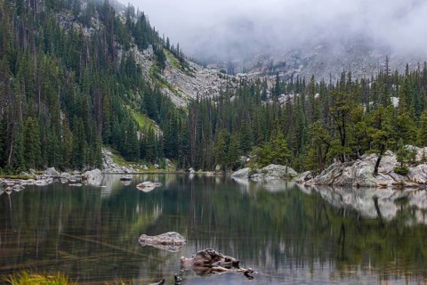 Misty atmosphere blankets a peaceful lake surrounded by dense forests and rugged mountains in the