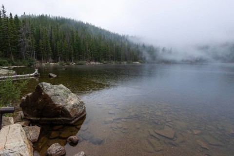 A foggy morning envelops a tranquil mountain lake surrounded by dense pine forests in Colorado,