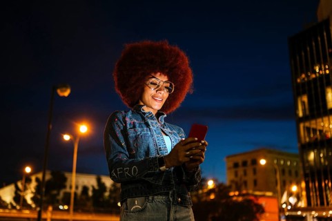 African descendent woman with afro hair, wearing glasses and a denim jacket, uses her smartphone on