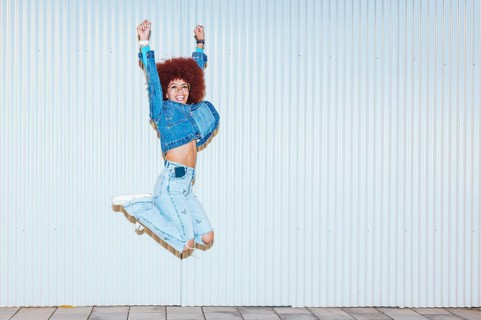 A joyful African descent woman with afro hair and fashionable denim outfit jumping energetically