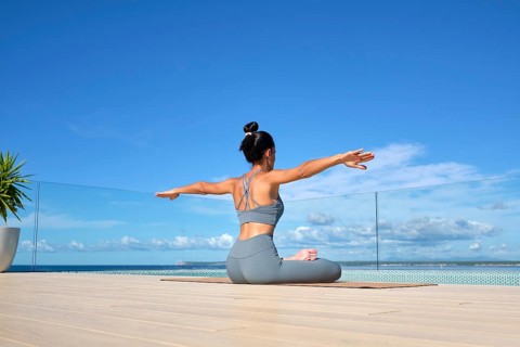 A woman in a gray yoga outfit practices yoga on a deck overlooking the ocean. Her arms are