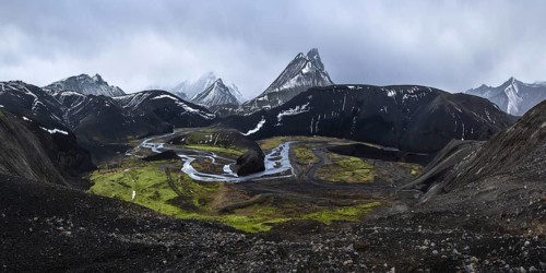 A striking view of black volcanic mountains partially covered in snow, with a winding river and