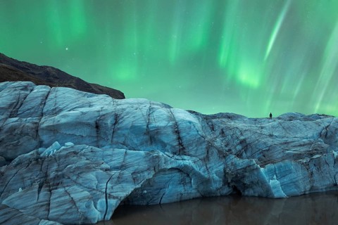A solitary figure stands atop a glacier in Iceland, gazing at the mesmerizing Northern Lights that