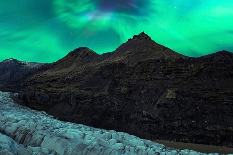 The Northern Lights cast a radiant glow over rugged mountains and a glacial landscape in Iceland,