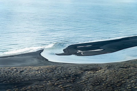 Aerial view of a winding Icelandic river meeting the sea. Black sand contrasts with the blue water,