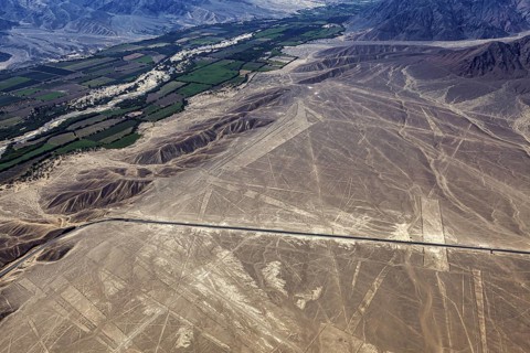 Aerial photo shows dry geoglyphic landscape next to green fields and a road, the geoglyphs and