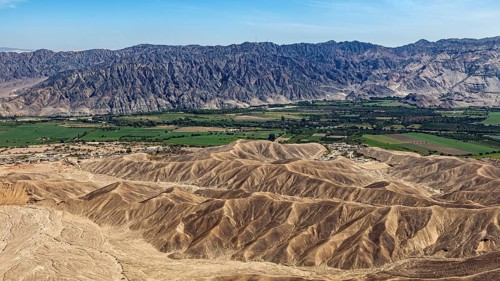 Contrasting landscape with desert in the foreground and green fields in front of a mountain chain,