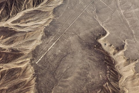 Sand-colored desert landscape with visible geoglyphs and linear patterns, surrounded by mountains,