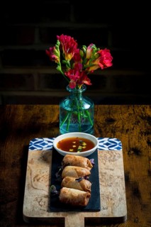 A wooden board holds crispy spring rolls beside a bowl of dipping sauce, set against a vibrant