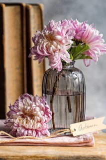 Soft pink chrysanthemums in a vintage glass vase beside rustic books and a tag reading Autumn