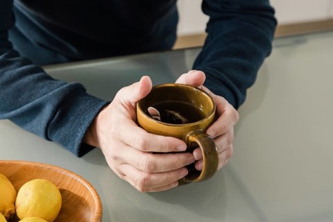A close-up of hands holding a brown mug of tea with a spoon inside, placed on a glass table A