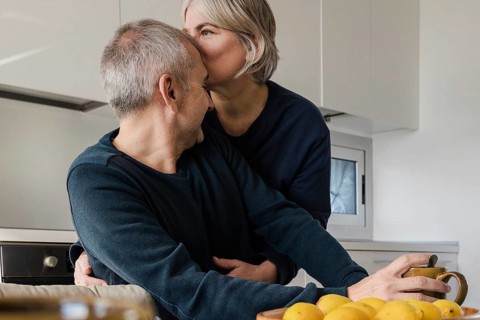 A senior couple enjoys a tender moment in their kitchen One person kisses the other on the head,