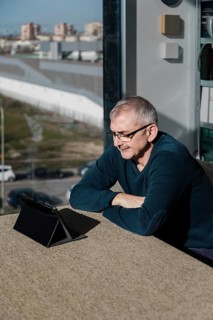 A senior man wearing glasses smiles as he engages with a tablet indoors Sunlight filters through