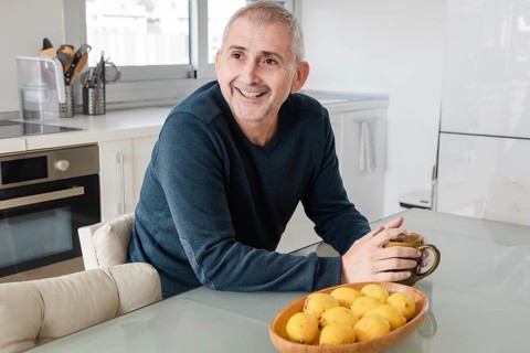 A cheerful senior man sits in a bright kitchen, holding a mug, with a basket of lemons nearby The