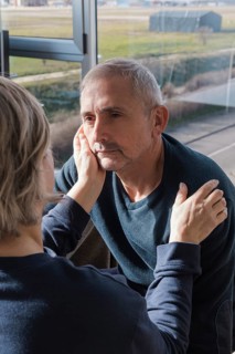 A senior couple shares a tender moment near a sunlit window, with one gently holding the other's