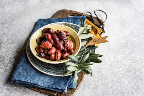 Ceramic bowl filled with Greek BBQ olives, served alongside fresh olive leaves and a rustic denim
