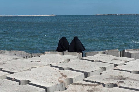 Two Muslim women wearing black abaya or chador sit on the concrete breakwater on the Mediterranean