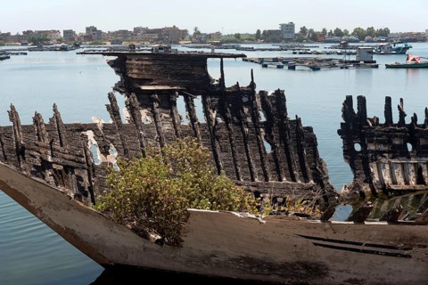 An abandoned burned out hull of a fishing boat with green plants growing inside it on the banks of