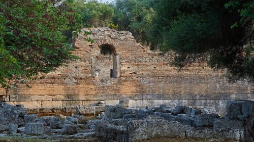 Palestra, red brick wall with an arch surrounded by vegetation and ruined architecture,