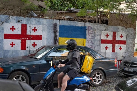 Tbilisi, Georgia. August 26th 2025. A motorcycle delivery driver rides past the flags of Georgia