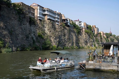 Tbilisi, Georgia. July 29th 2025. A tourist river cruise passes under building sitting precariously
