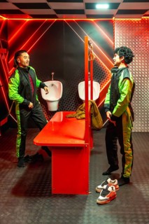 Two young men in green and black jumpsuits chat in a red neon locker room, gearing up and bonding