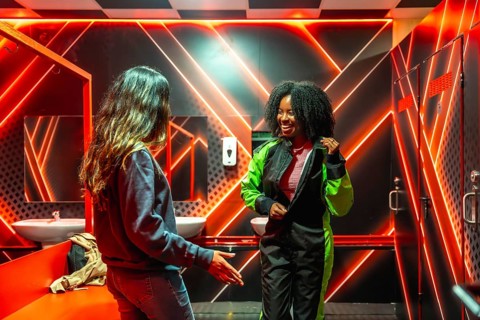 Two cheerful women are preparing for a gokarting race in a neon lit locker room, with one friend