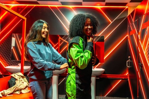 Two diverse friends laughing and helping each other put on racing suits in a changing room with