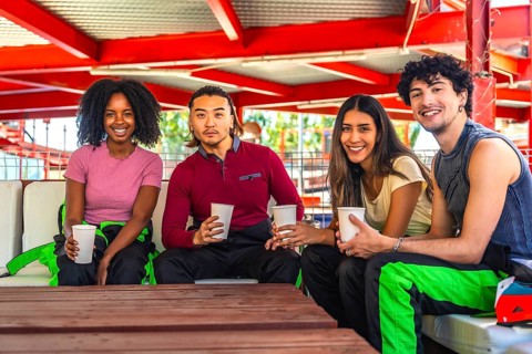 Multiethnic group of young adults in racing suits relaxing at a table with drinks, smiling and