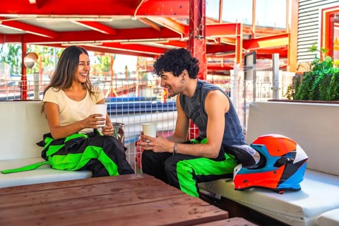 Young couple in racing suits sharing coffee and laughter at a trackside table after karting,