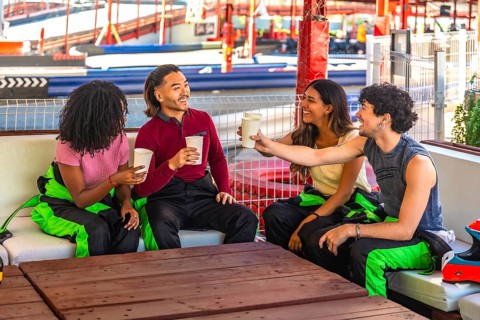 Group of diverse young adults wearing racing suits sitting on a couch, laughing and toasting each