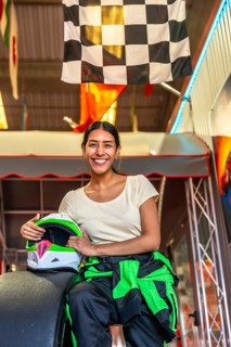 Young woman in racing suit sits in a go kart holding her helmet beneath a checkered flag, smiling