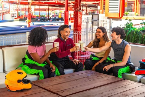 Four diverse young friends in racing suits making a celebratory toast with drinks at a wooden table