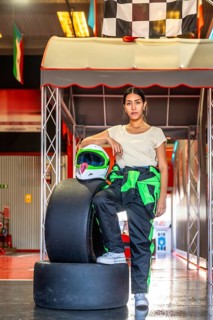 Woman wearing a racing suit and helmet standing confidently by tires and checkered flags at an