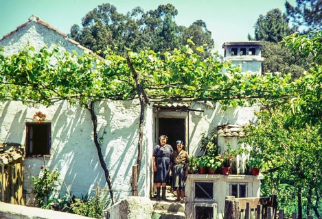 Two women standing outside a traditional rural house with grapevines growing in the garden,