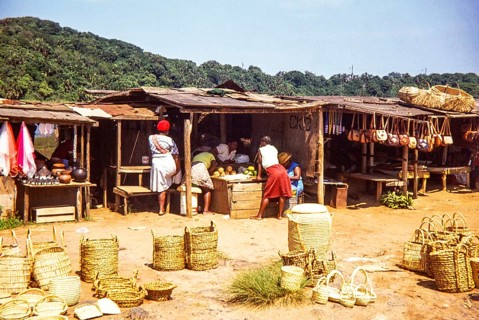 Woman at local market selling fruit and handicrafts including baskets, leather handbags and