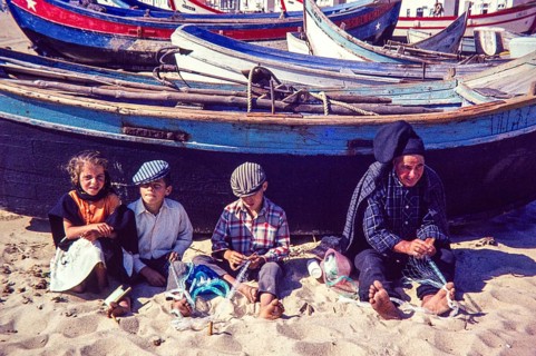 Fisherman and children repairing nets on beach by traditional fishing boats, Nazaré, Portugal,