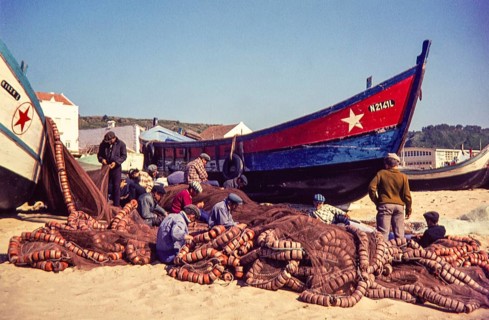 Fishermen repairing nets on beach by traditional fishing boats, Nazaré, Portugal, Europe 1968