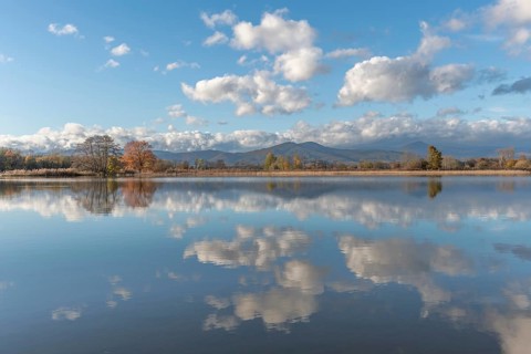 The river flows flat and reflects clouds and mountains under a clear sky. Autumn trees draw the