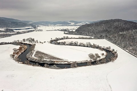River flows in a loop through snow-covered fields, framed by hills and trees under cloudy skies,