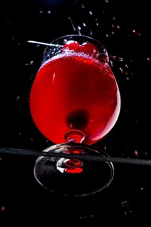 Sparkling red drink with cherries in a cognac glass on black background, view from below